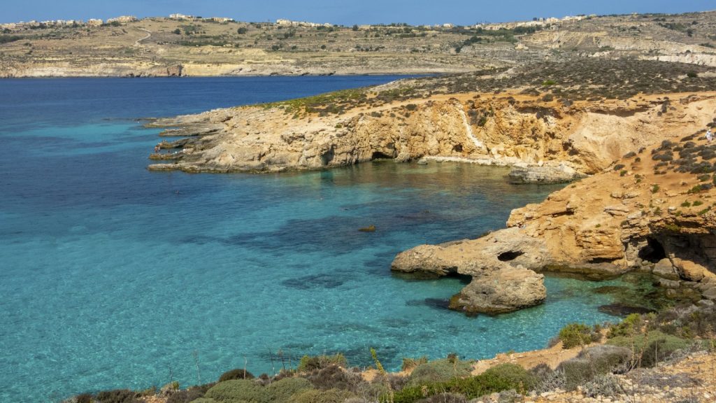 Comino sziklás partja kristálytiszta türkiz vízzel, háttérben Gozo szigete