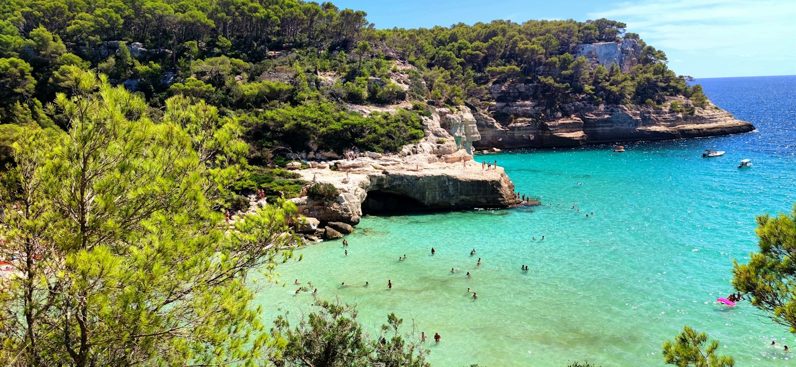 Türkiz víz és fenyves mészkősziklák Cala Mitjana öblében, Menorca déli partján